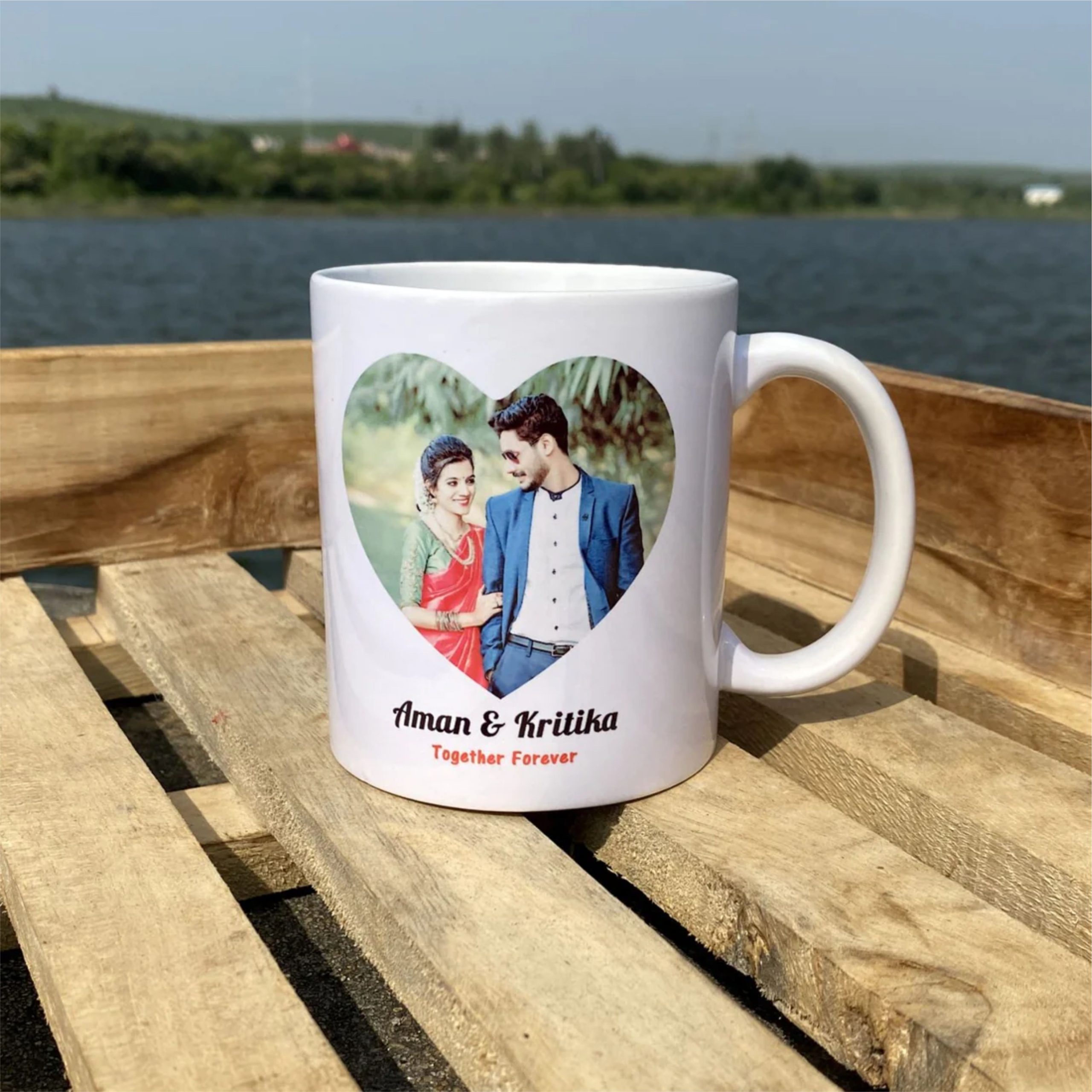 Personalized white ceramic couple mug featuring a custom photo inside a heart design, names printed below, and a calendar highlighting a special date. The customized coffee mugs are displayed on a wooden table beside a scenic lakeside background, making a perfect anniversary or romantic gift.